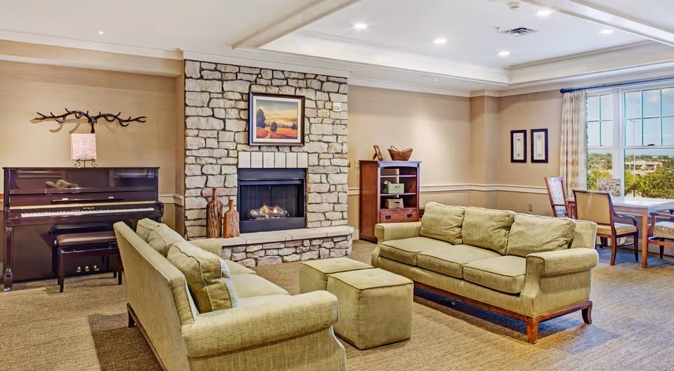 A cozy communal living room with two green sofas and an ottoman facing a stone fireplace, a piano on the left, and a dining table by a window.