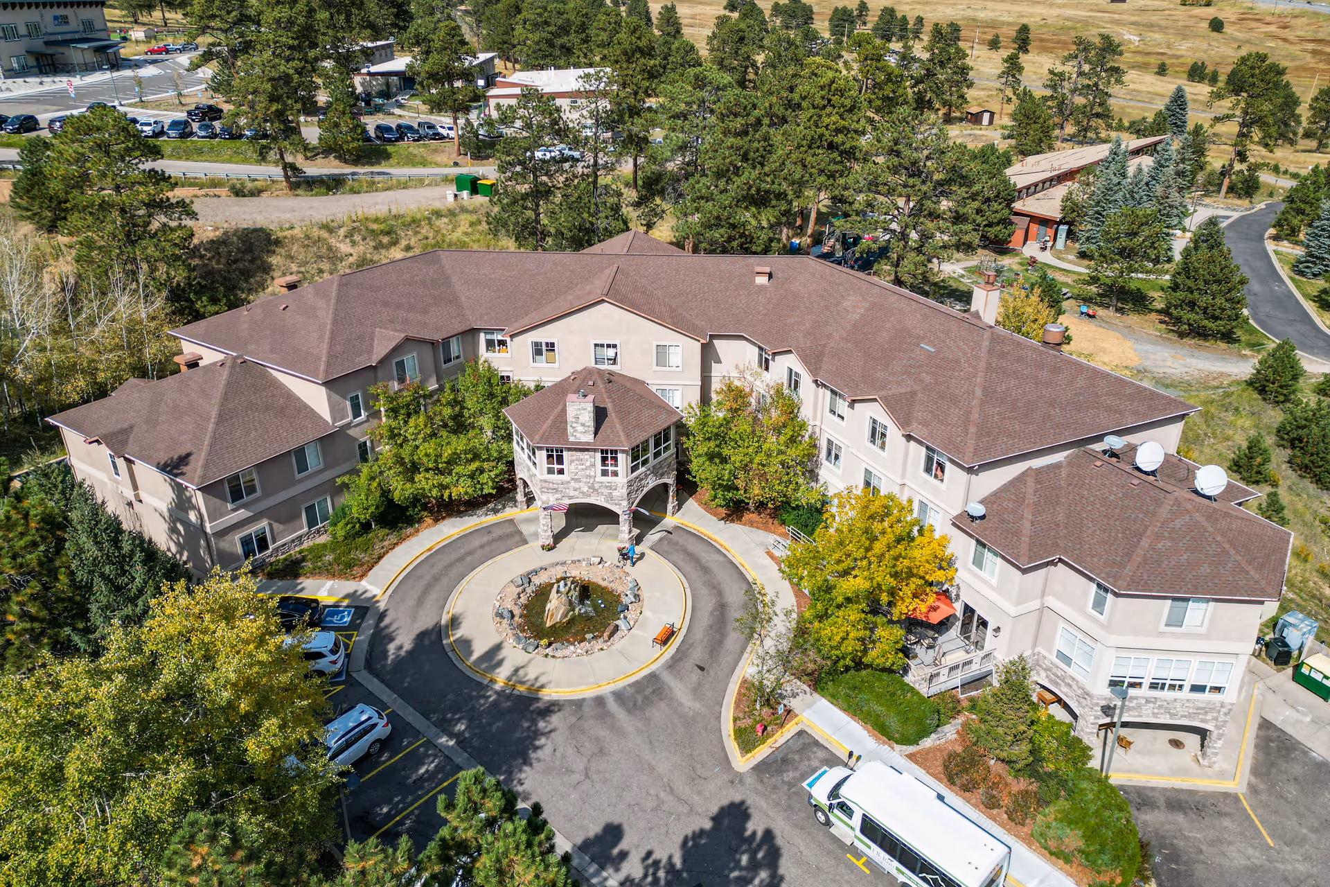Aerial view of a large assisted living facility named Elk Run Assisted Living, featuring a U-shaped building with a brown roof, surrounded by trees and greenery. There is a circular driveway with a small stone gazebo and a rock fountain in the center. Several vehicles are parked around the building, and the surrounding area includes roads and open land.