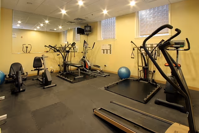 Interior view of a fitness room with various exercise equipment including a treadmill, stationary bikes, elliptical machines, and a blue exercise ball. The room has yellow walls, a black rubber floor, and windows with glass blocks letting in natural light.