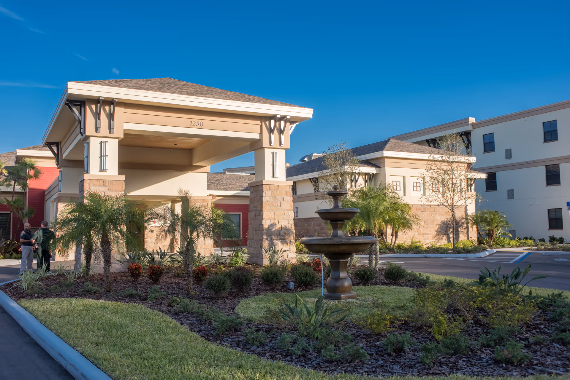 Exterior view of The Fountains of Hope senior living facility entrance with a covered drop-off area, landscaped garden with palm trees and shrubs, and a tiered water fountain in the foreground under a clear blue sky.