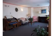 Small living room/common area with upholstered chairs, a floral sofa, an organ, coffee table, plants, and framed art on the wall.