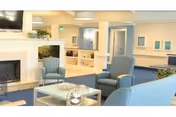A bright and spacious living room area with blue upholstered chairs and sofas arranged around a glass coffee table. There is a white fireplace on the left side and built-in shelving with decorative items. The walls are light-colored with framed artwork, and the floor is carpeted in blue.