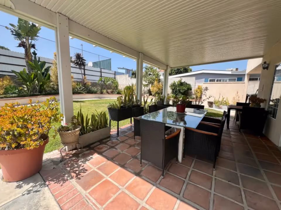 Covered outdoor patio area with a glass-top table surrounded by black wicker chairs. Several potted plants are placed around the patio, and a green lawn with various plants and trees is visible beyond the patio. The sky is clear and blue.
