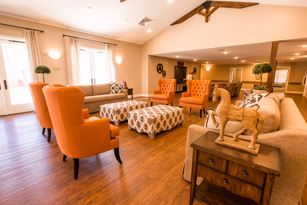 A spacious and well-lit living room area in a senior living facility featuring multiple orange armchairs, beige sofas, patterned ottomans, wooden flooring, and decorative elements including a wooden horse sculpture on a side table. Large windows with curtains allow natural light to fill the room.