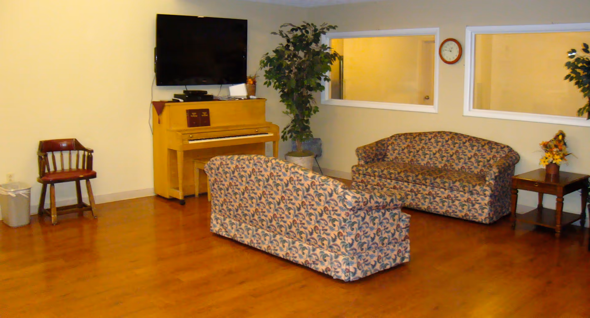 A common room with patterned sofas, a piano under a wall-mounted TV, a small table with flowers, and wood flooring.