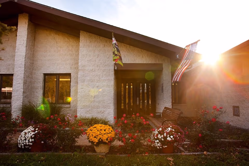 Front entrance of a single-story building with flowers, a bench, flags and sunlight flaring at sunset.