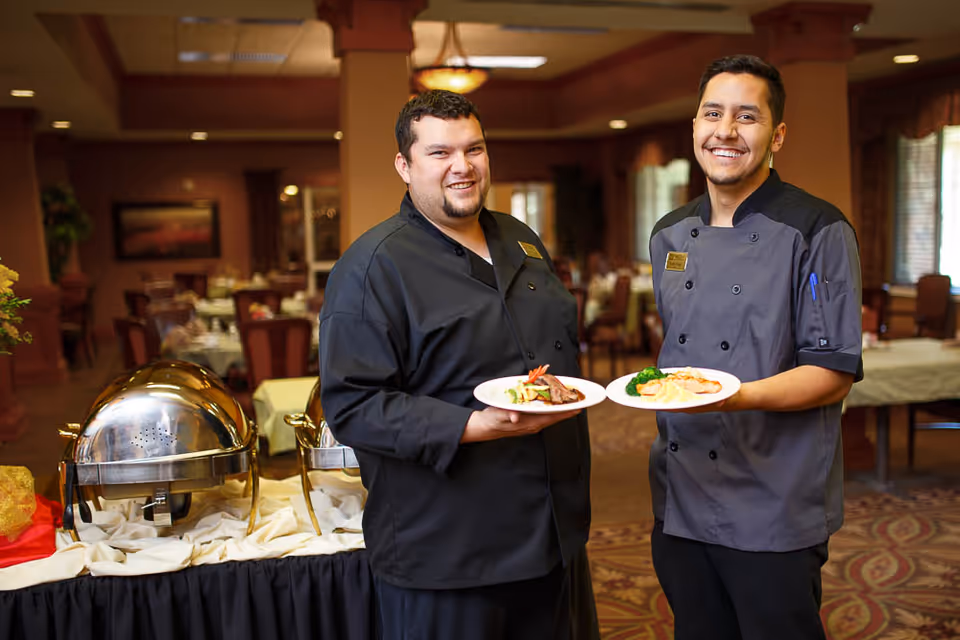 Two smiling chefs standing in a dining room holding plates of food, with buffet chafing dishes and tables set for dining in the background.