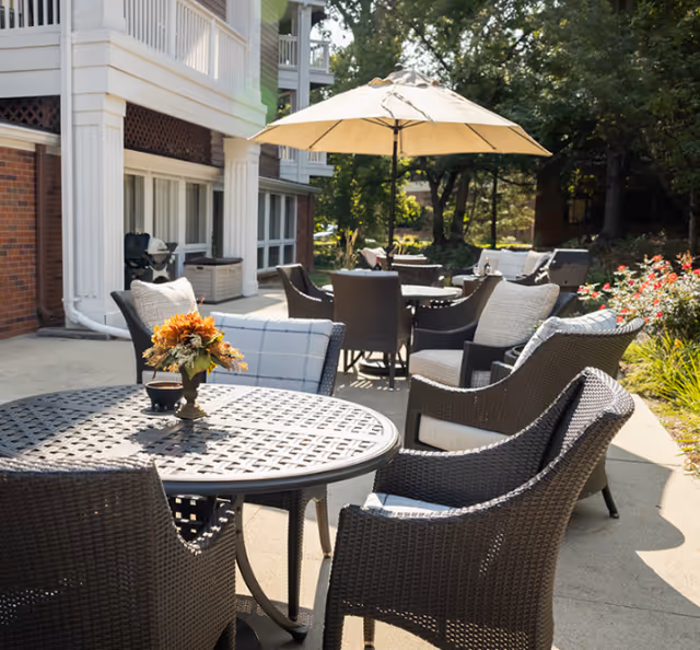 Outdoor patio area at Bloomfield facility with several dark wicker chairs and round tables. One table has a flower arrangement, and another table has a large beige umbrella providing shade. The patio is adjacent to a brick building with white columns and surrounded by greenery and trees.
