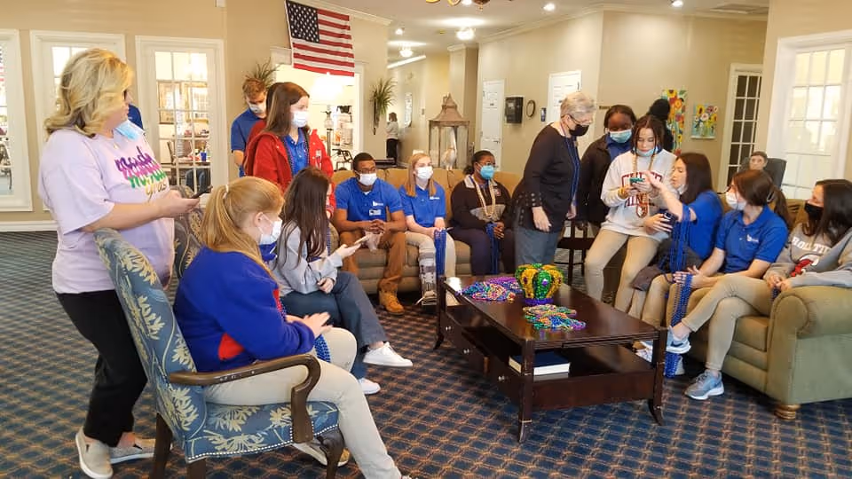 A group of people, mostly wearing blue uniforms and face masks, are gathered in a well-lit living room with patterned carpet and beige walls. Some are seated on chairs and sofas around a wooden coffee table decorated with colorful Mardi Gras beads and a crown. One woman stands near the left side of the image, and others are engaged in conversation or looking at their phones.