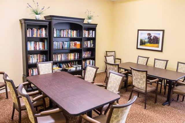 A room with two dark wooden tables surrounded by cushioned chairs with patterned upholstery. Against the wall is a large dark wooden bookshelf filled with books and decorated with two small potted plants on top. The walls are painted light yellow, and there is a framed picture of a landscape hanging on the wall.