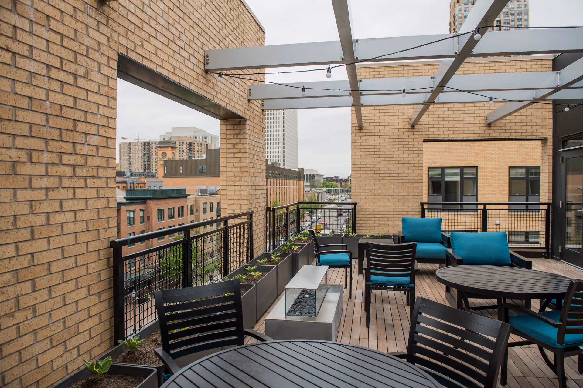 A rooftop patio with round tables, chairs, teal-cushioned lounge seating, planters, and a view of surrounding city buildings.