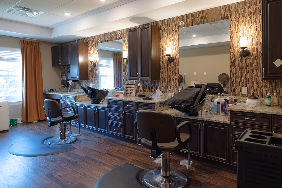 Interior view of a salon area with two salon chairs in front of a long counter with sinks and mirrors. The counter has dark wood cabinets and a mosaic tile backsplash. There are wall-mounted lights above the mirrors and a window with brown curtains on the left side.