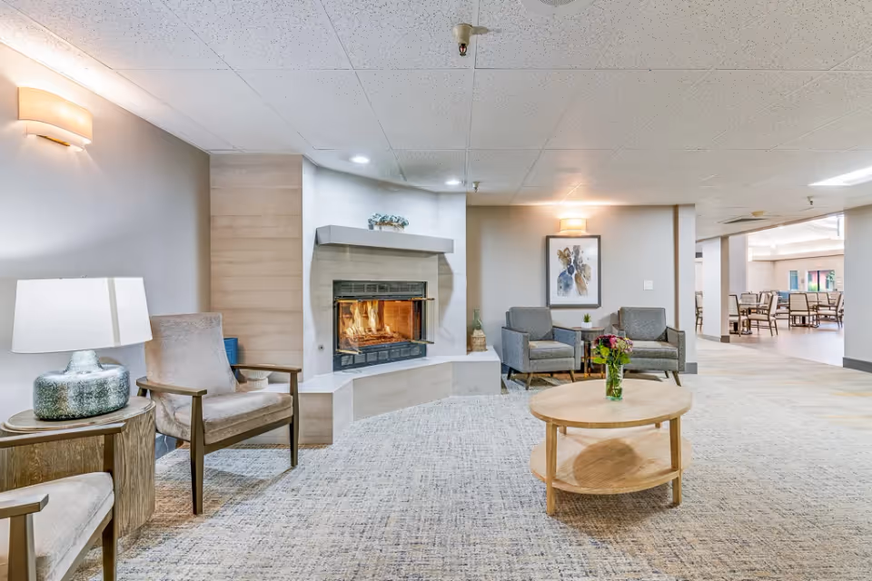 A cozy living room area with a lit fireplace in the corner, surrounded by four armchairs and a round wooden coffee table with a vase of flowers. The room has soft lighting from wall sconces and a table lamp, with a framed artwork on the wall and an open view into a dining area with tables and chairs.