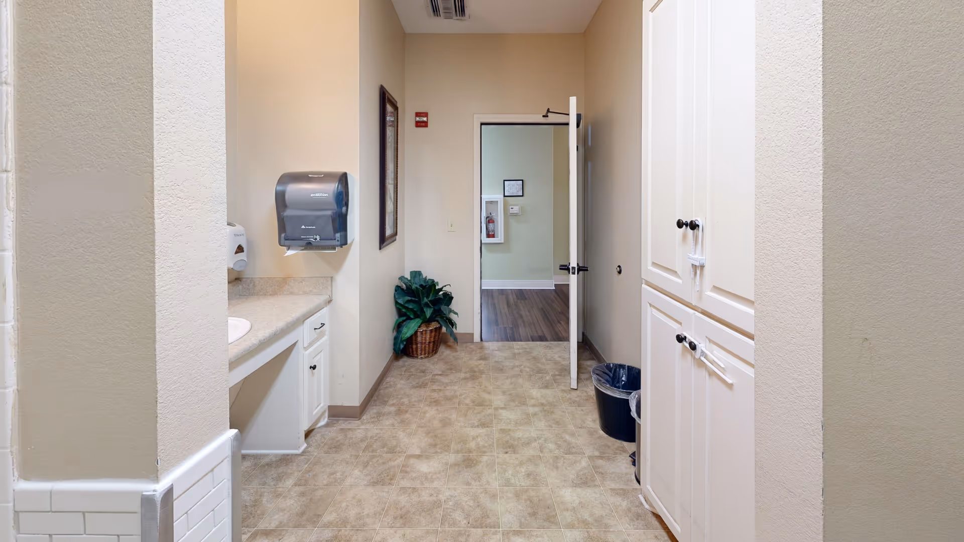 Interior corridor with a countertop sink, wall-mounted paper towel dispenser, tall cabinets, trash can, potted plant, and an open door leading to another room.