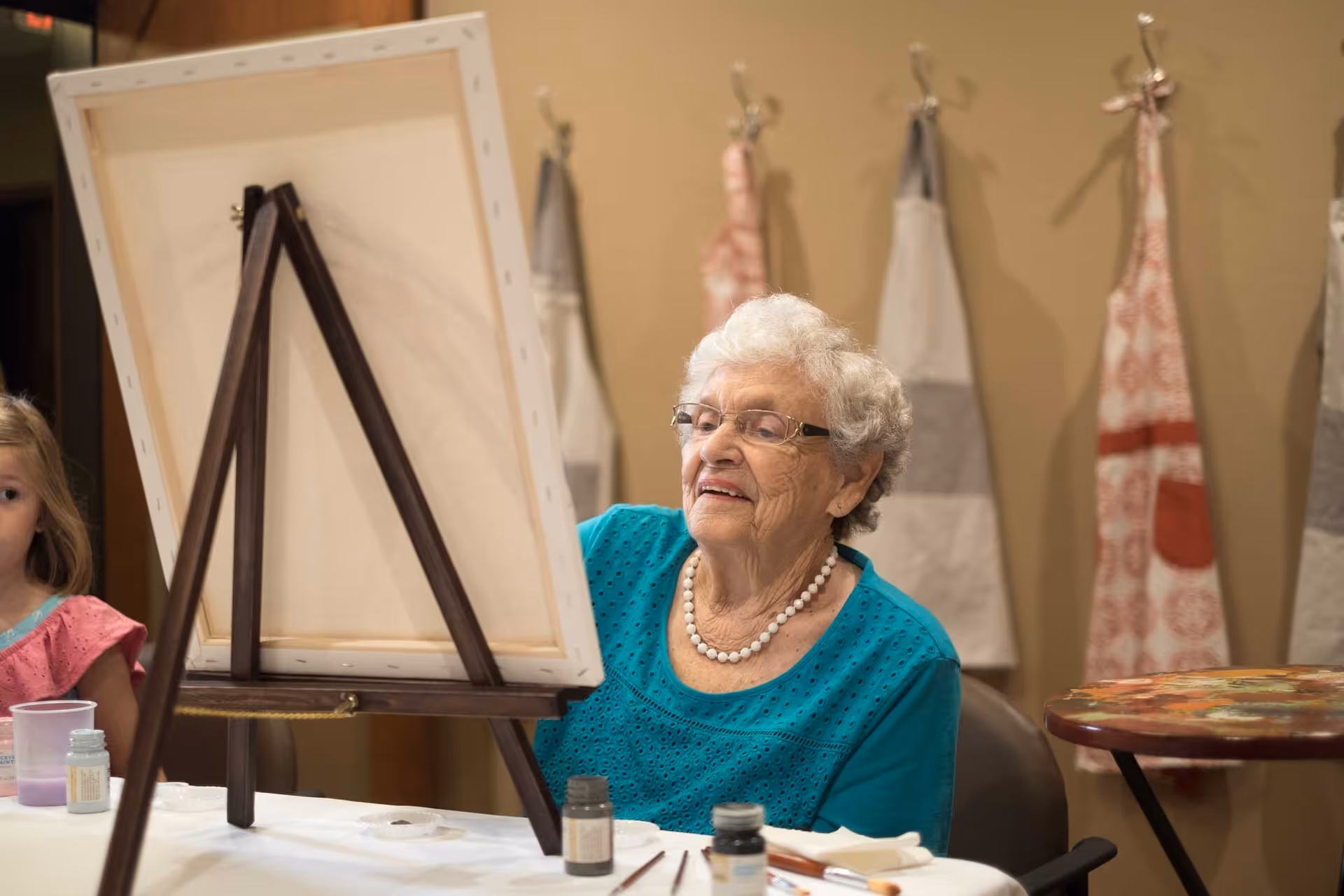 An elderly woman wearing glasses and a turquoise top is sitting at a table painting on a canvas set on an easel. She is smiling and appears focused on her artwork. In the background, aprons are hanging on hooks on the wall. A young girl is partially visible on the left side of the image.