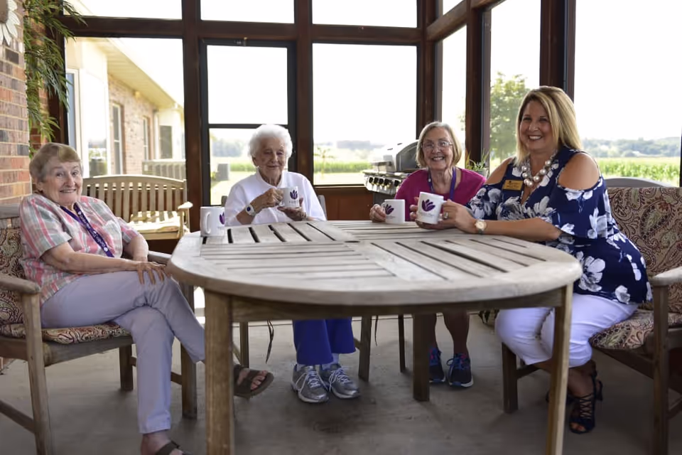Four women sitting around a wooden table on a screened porch, holding white mugs and smiling. The porch overlooks a green outdoor area with fields in the background.