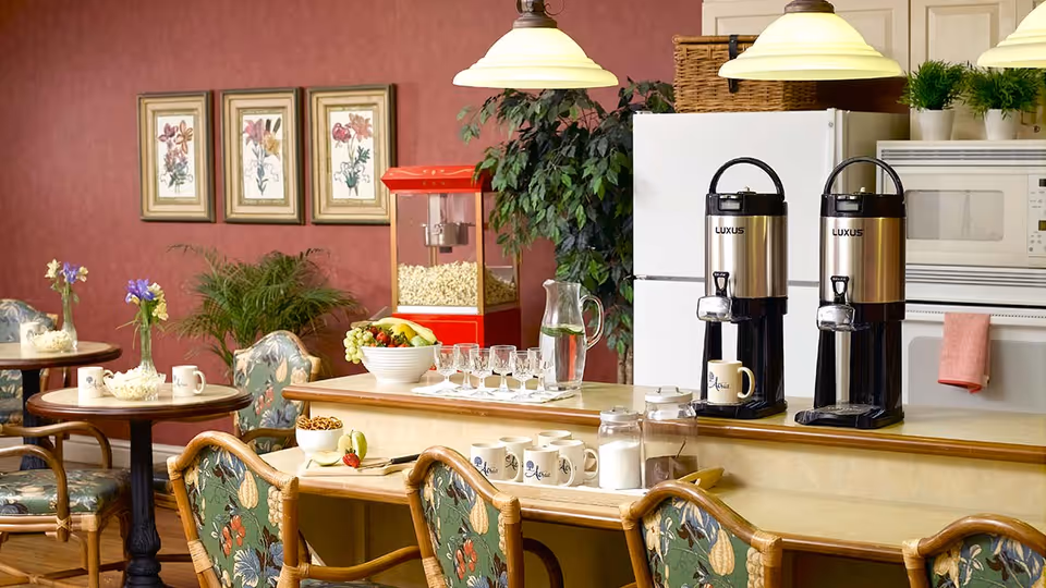 A cozy dining area with small round tables and floral cushioned chairs. On the counter, there are two large coffee dispensers, a popcorn machine, a bowl of fruit, glassware, and mugs. The background features a refrigerator, microwave, and framed floral artwork on a red wall.