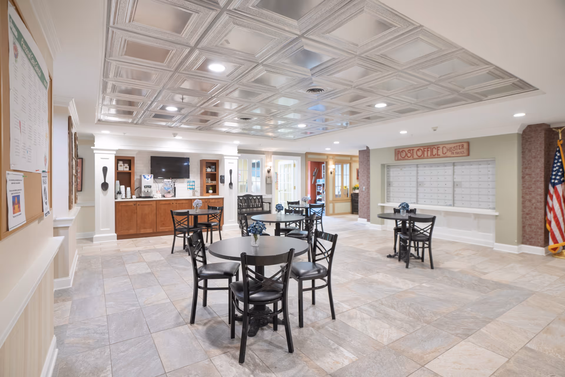 A spacious common area with several round tables and black chairs arranged for seating. The room features a decorative ceiling with recessed lighting, a bulletin board on the left wall, a coffee station with a TV mounted above it, and a wall of mailboxes labeled 'Post Office Chester'. An American flag stands in the corner on the right side.