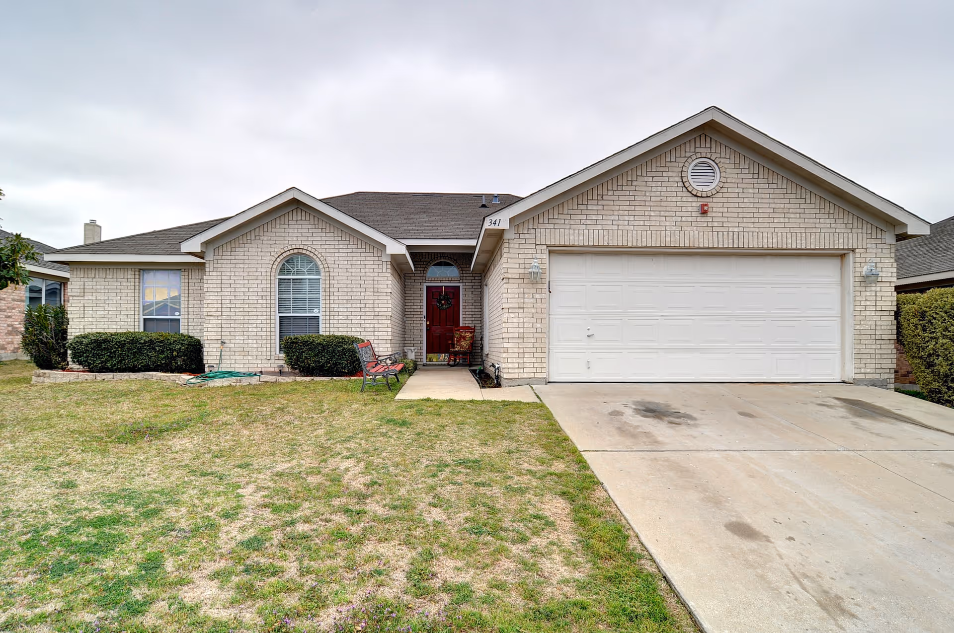 Front exterior of a single-story brick house with a two-car garage, driveway, lawn, and a walkway to a red front door.