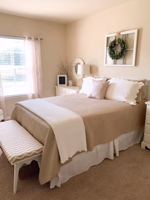 A cozy bedroom with a neatly made bed featuring beige and white bedding, decorative pillows, and a white throw blanket. A cushioned bench with a chevron pattern sits at the foot of the bed. There is a window with sheer curtains allowing natural light to fill the room. A nightstand with a lamp, a small clock, and decorative items is beside the bed. A framed window pane with a green wreath hangs on the wall above the bed.