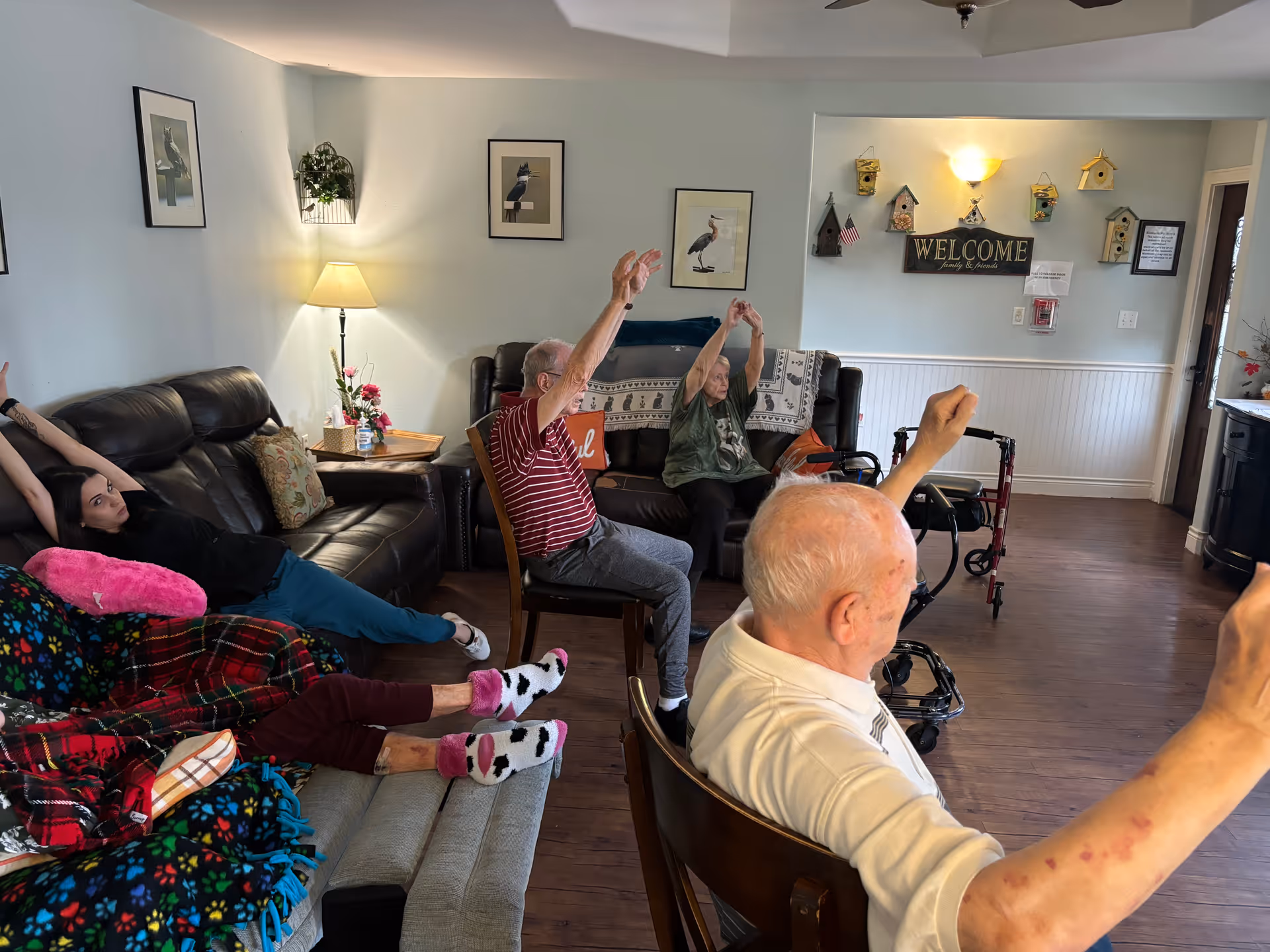 A group of elderly people and a younger woman participating in a seated exercise session in a cozy living room with leather sofas, framed bird pictures on the wall, a lamp, and decorative birdhouses with a welcome sign.