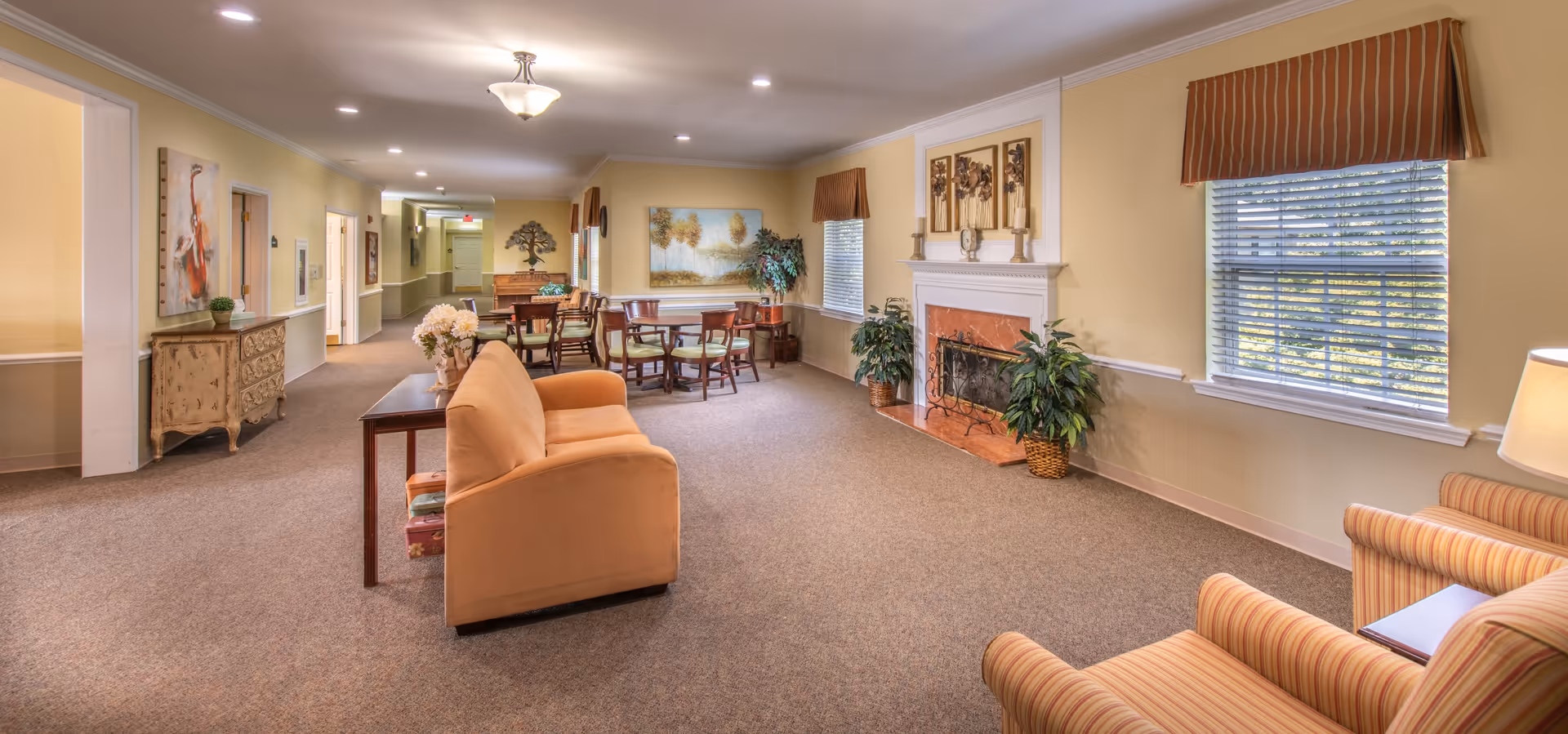 Spacious furnished common room with sofas, armchairs, a fireplace and dining tables in a senior living facility.