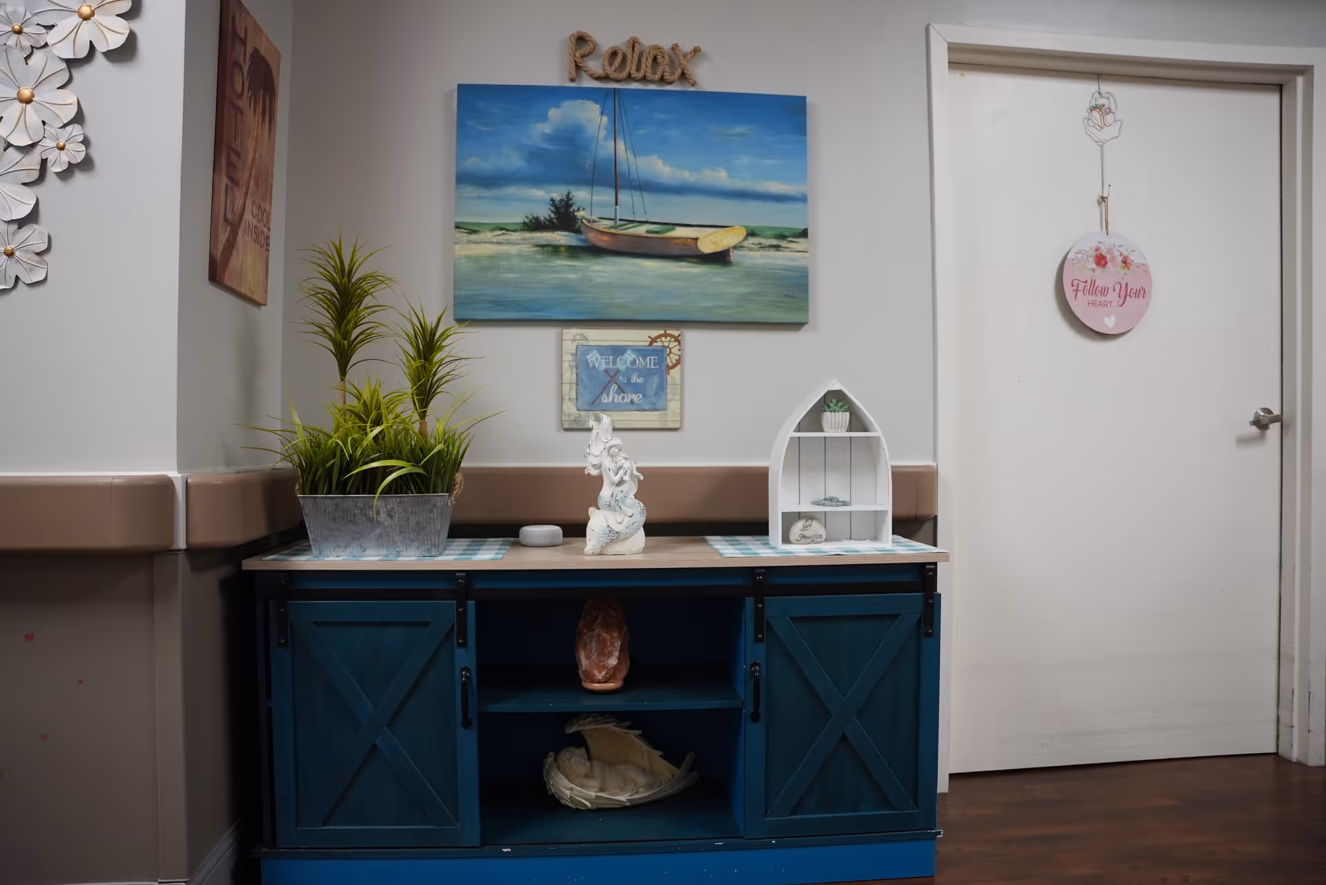 A decorative corner in a facility with a blue cabinet featuring sliding barn doors. On top of the cabinet are green plants in a metal container, a white seahorse figurine, and a small white shelf shaped like a boat holding a small plant and seashells. Above the cabinet, there is a painting of a sailboat on water with the word 'Relax' above it, and a small sign that says 'Welcome to the shore'. To the right, a white door has a hanging round sign that reads 'Follow Your Heart'. The walls are painted light gray with a brown chair rail, and the floor is dark wood.
