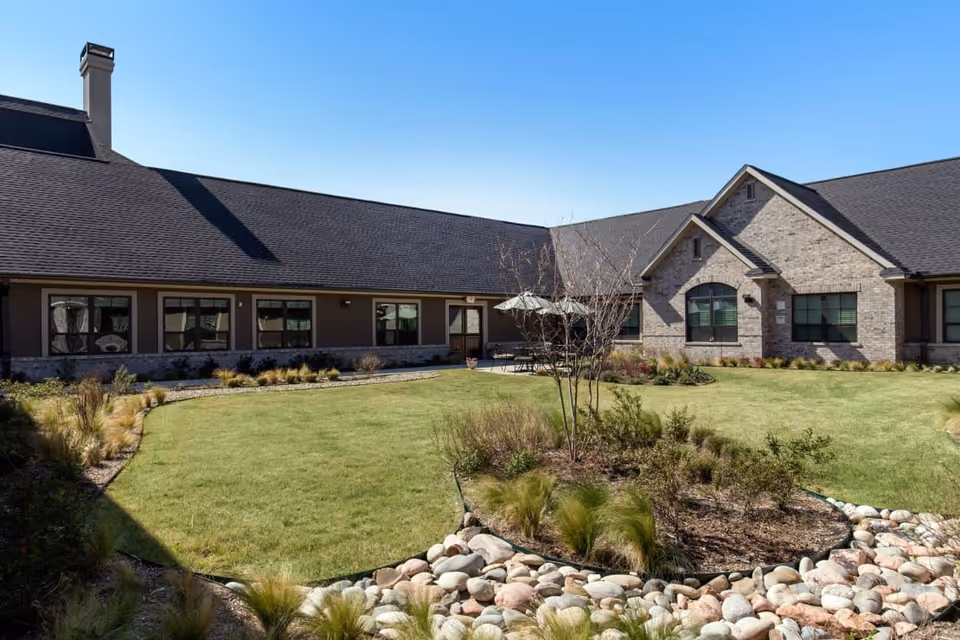 Outdoor courtyard area of Briarview Senior Living with a well-maintained lawn, landscaped plants, and a stone border. The building surrounding the courtyard has a dark roof, brick and siding exterior, and multiple windows. There are patio tables with umbrellas near the building.
