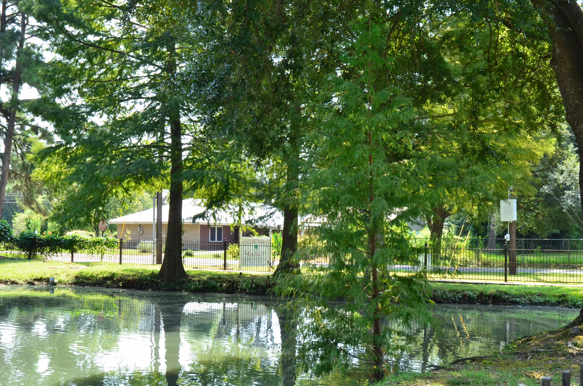 A reflective pond in front of large trees with a fence and a low brick building visible through the foliage.