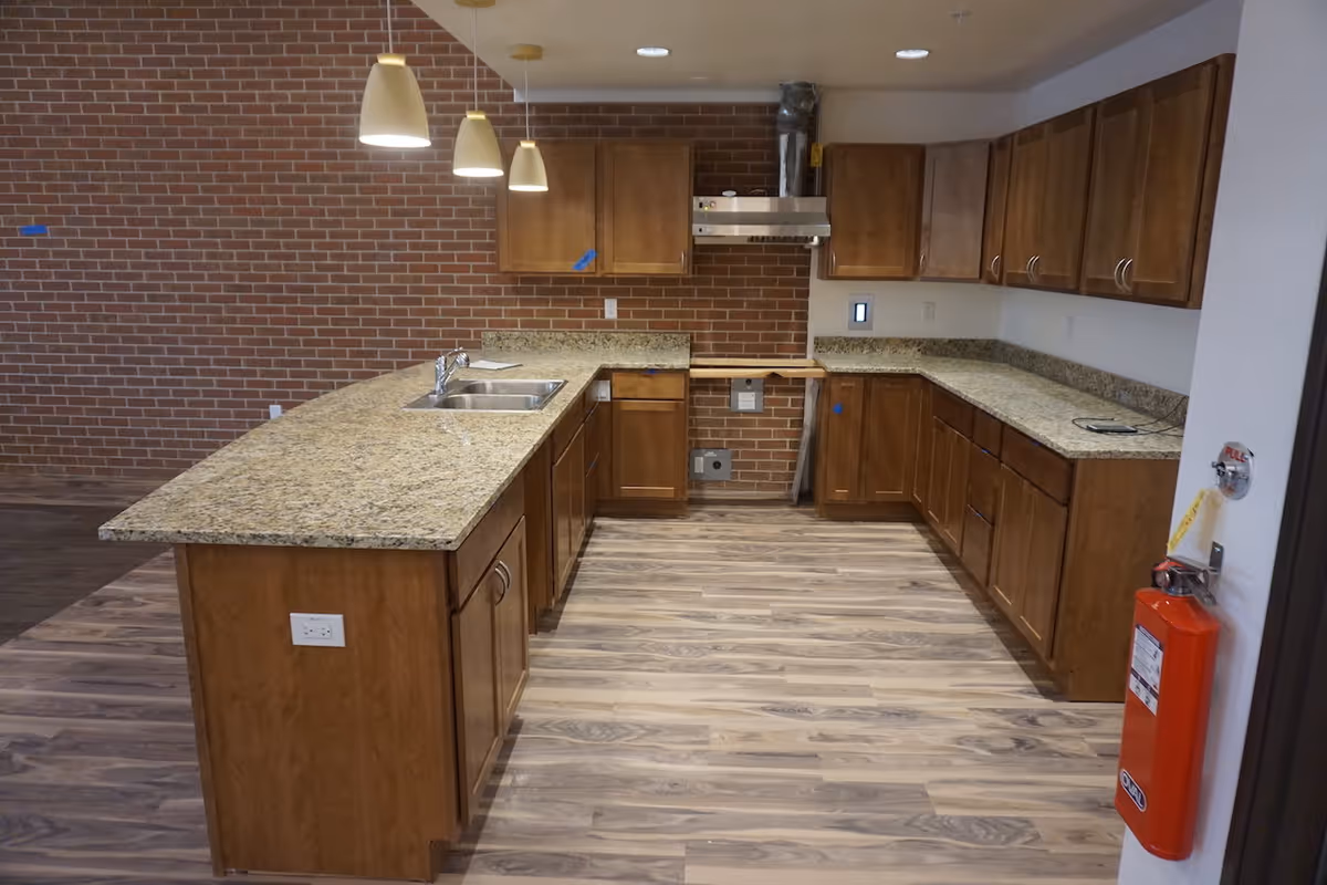 A spacious kitchen with wooden cabinets and granite countertops. The kitchen features a double sink on the island, three pendant lights hanging above the island, a stainless steel range hood, and a brick wall backdrop. The floor is covered with wood-patterned vinyl or laminate. A red fire extinguisher is mounted on the right wall near the entrance.