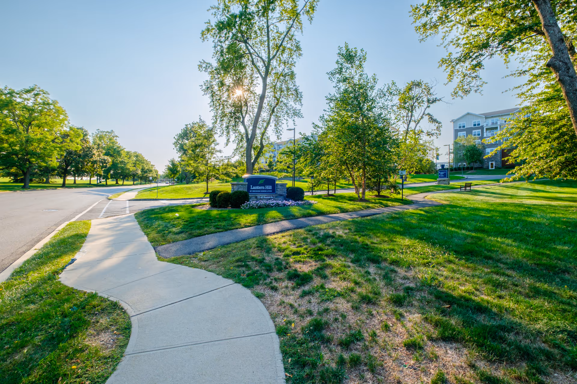 A sunny outdoor scene at Lantern Hill featuring a curved sidewalk, green grass, trees, and a sign with the facility's name. Residential buildings are visible in the background.