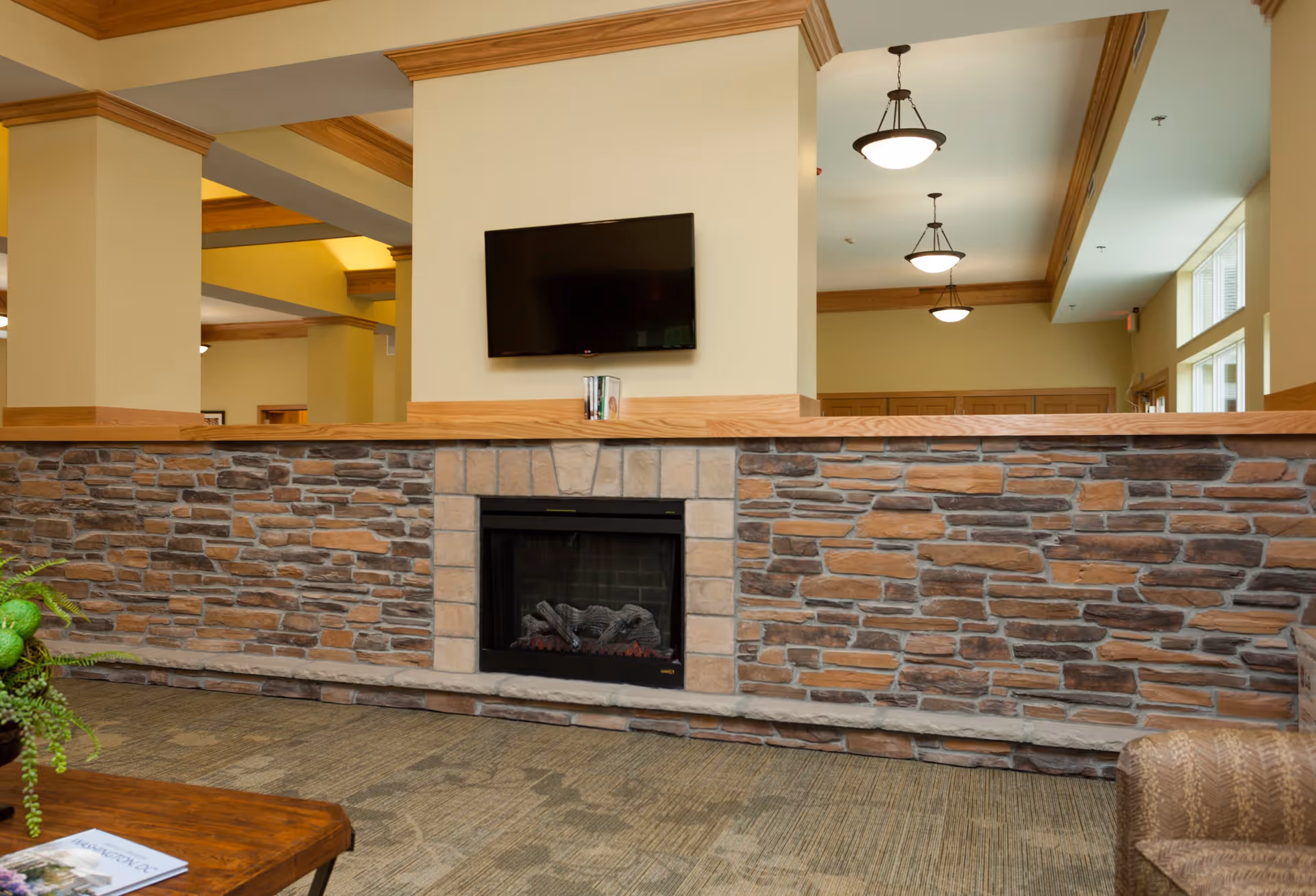 Interior view of a senior living facility lounge area featuring a stone fireplace with a wooden mantle, a flat-screen TV mounted above it, beige walls with wooden trim, carpeted floor, and ceiling lights. A small part of a wooden table with a plant and a magazine is visible in the foreground.