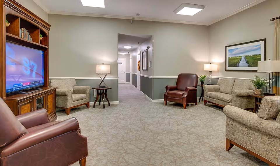 A cozy senior living facility common area with beige patterned carpet, light gray walls with white trim, and soft lighting from table lamps. The room features a wooden entertainment center with a TV, two brown leather armchairs, two beige upholstered armchairs, a beige sofa, side tables with lamps and plants, and a framed landscape picture on the wall. A hallway is visible in the background.