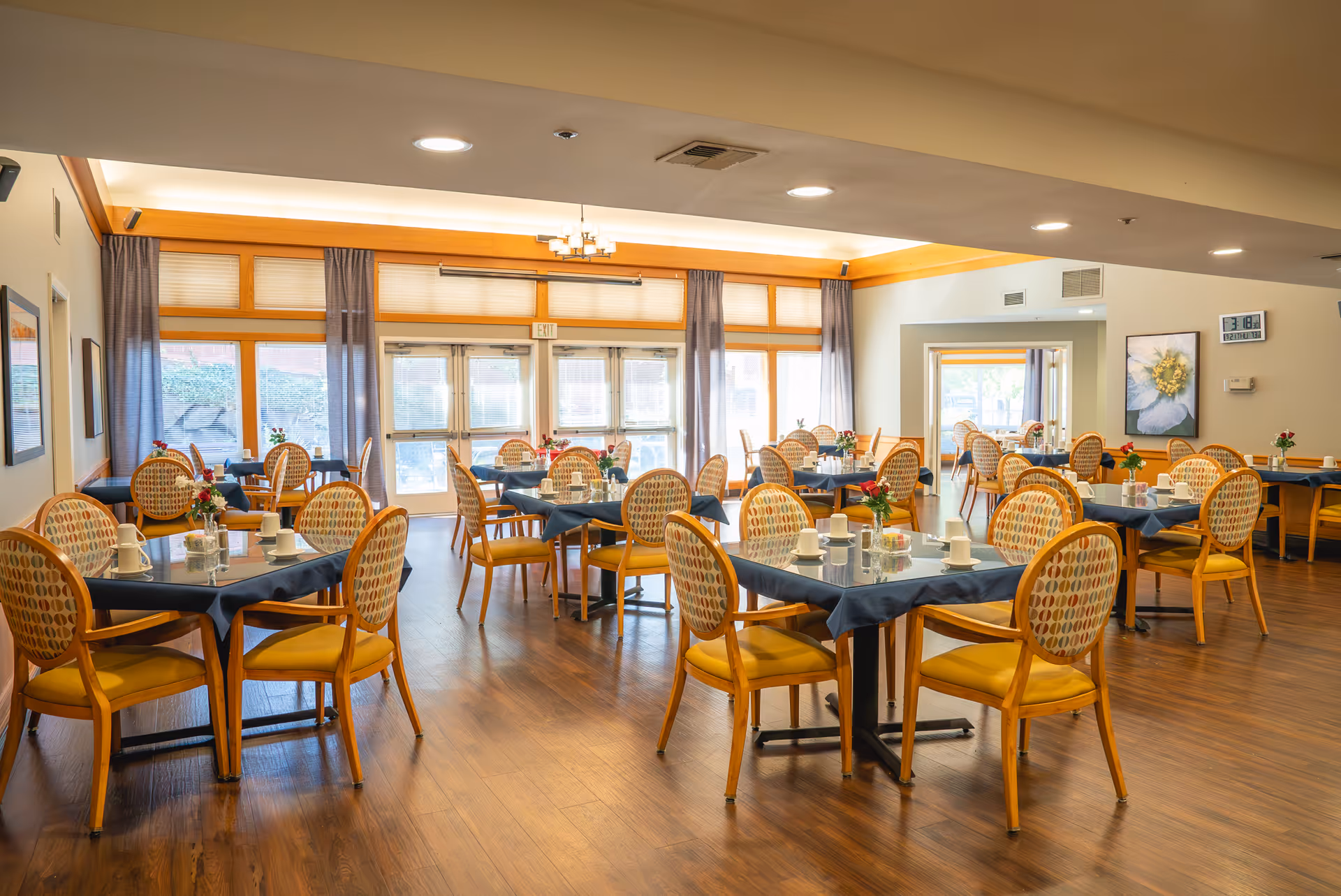 A bright and spacious dining room with multiple round tables covered with dark tablecloths, each surrounded by yellow cushioned chairs with patterned backs. The tables are set with white cups, saucers, and small flower arrangements. Large windows with sheer curtains allow natural light to fill the room, and the floor is made of polished wood.