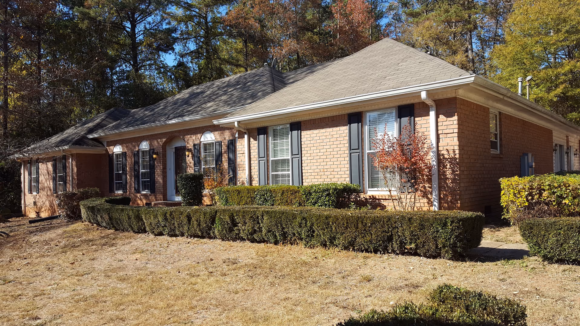 Exterior view of a single-story brick building with a gray shingled roof, black window shutters, and a white front door. The building is surrounded by trimmed hedges and bushes, with trees in the background under a clear blue sky.