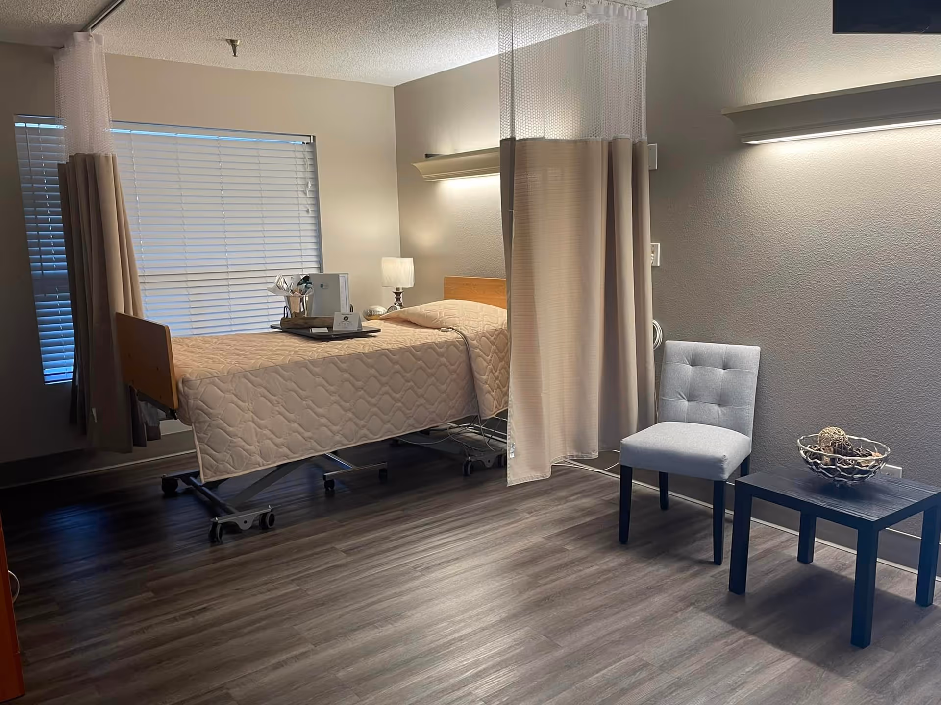 A nursing facility room with a hospital bed covered in a beige quilted blanket, a small bedside table with a lamp, and a tray with items on the bed. There is a privacy curtain partially drawn around the bed. Next to the bed is a gray upholstered chair and a small black table with a decorative bowl. The room has wood-look flooring and a window with closed blinds.