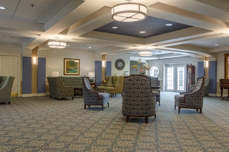 Spacious furnished common lounge with patterned carpet, armchairs and sofas, overhead light fixtures, and double doors at the far end.