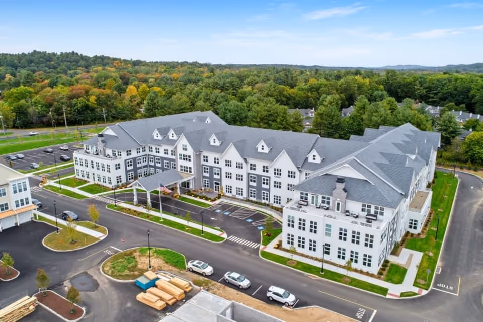 Aerial view of a large, modern senior living facility named Skybrooke at Simsbury, surrounded by trees and greenery. The building has multiple floors with gray roofs and white exterior walls. There is a parking lot with several cars and landscaped areas around the building.