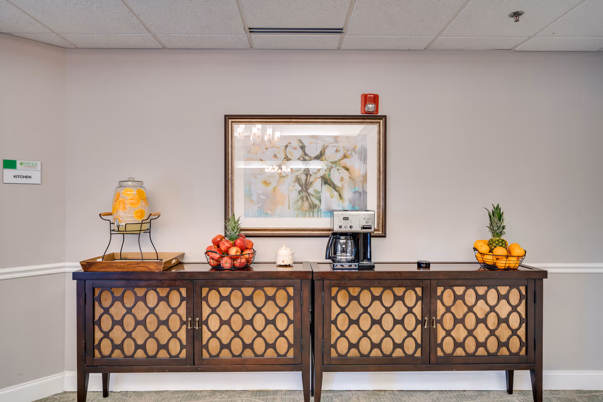 A wooden buffet cabinet against a wall topped with a coffee maker, a glass beverage dispenser, bowls of fruit, and a framed painting.