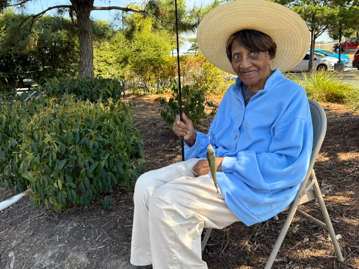 An elderly woman wearing a large straw hat and a light blue jacket sits on a folding chair outdoors, holding a fishing rod with a small fish caught on the line. She is surrounded by greenery and trees, with parked cars visible in the background.