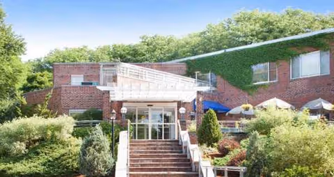 Exterior brick building entrance with stairs, greenery, and outdoor seating.