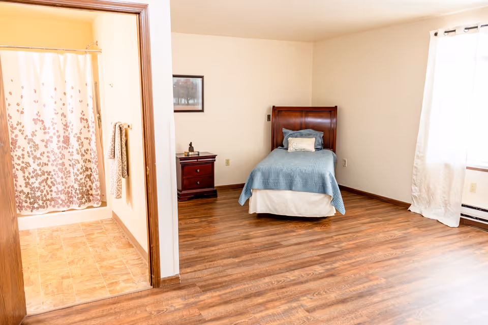 A simple bedroom with a single bed and nightstand, hardwood floors, a window with curtains, and an adjoining bathroom with a shower.