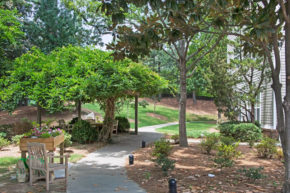 A shaded paved path leading past a pergola-covered seating area with benches, planters, and landscaped trees beside a building.