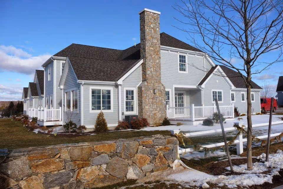 Exterior view of a light gray two-story residential building with white trim and a stone chimney. The building has multiple windows and a white porch railing. There is a stone wall and some small trees in the foreground with patches of snow on the ground under a clear blue sky.