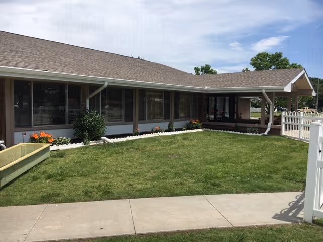 Exterior view of a single-story building with a sloped roof, large windows, and a well-maintained lawn with flower beds. A concrete sidewalk runs along the front, and a white fence is visible on the right side under a partly cloudy sky.