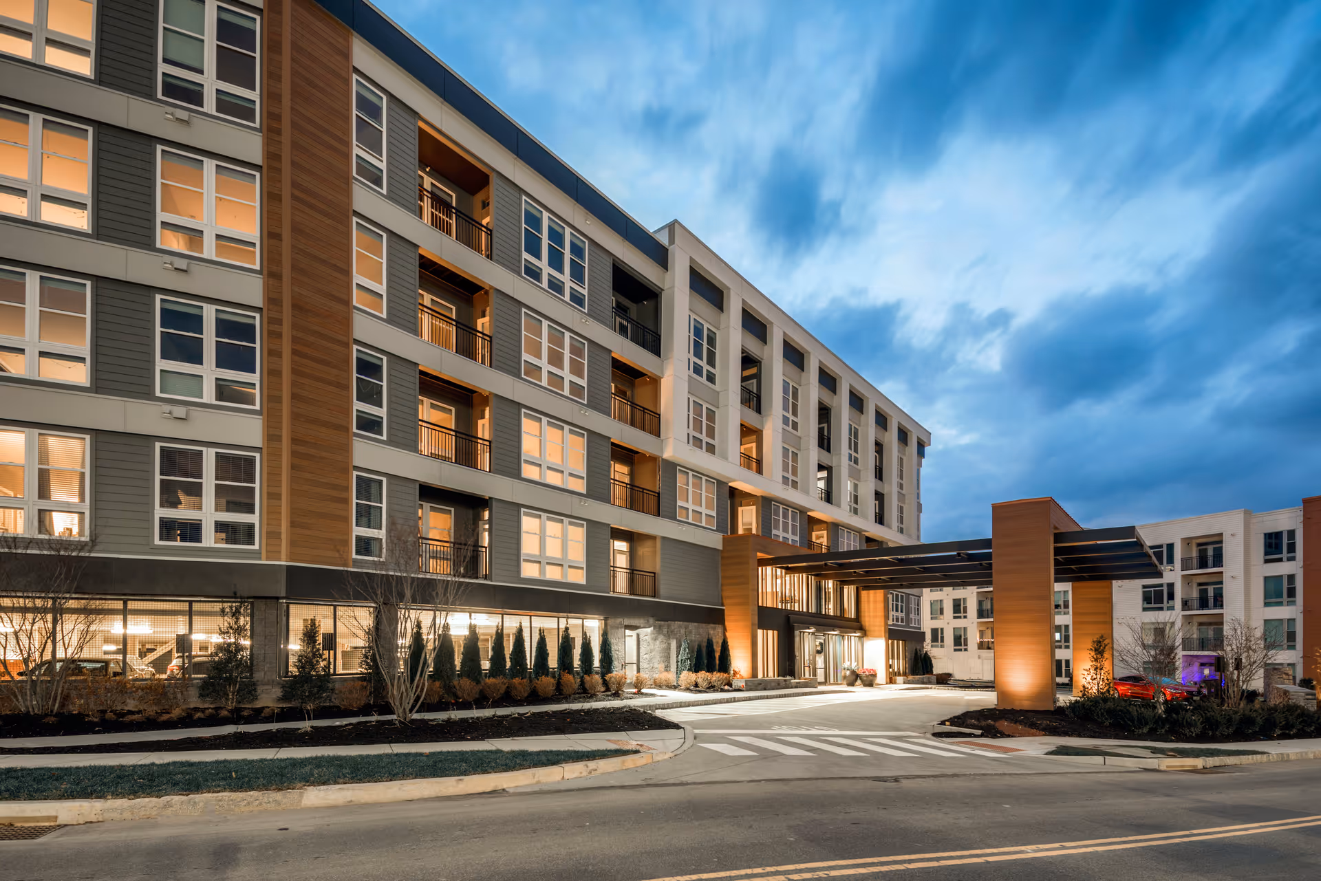 Exterior view of a modern multi-story senior living facility named Canvas Valley Forge during dusk, with warm lights glowing from the windows and a covered entrance area with pillars.