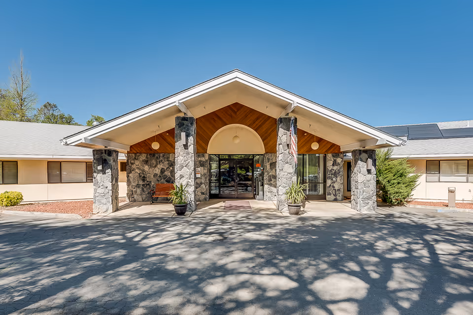 Front entrance of a one-story nursing facility with a covered porte-cochere supported by stone columns, potted plants, a bench, and glass doors under a clear blue sky.