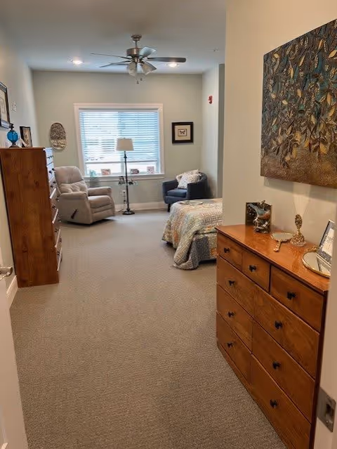 A cozy bedroom with a bed covered in a quilt, a reclining chair, a small armchair, a floor lamp near a window with blinds, and wooden dressers with decorative items and artwork on the walls.