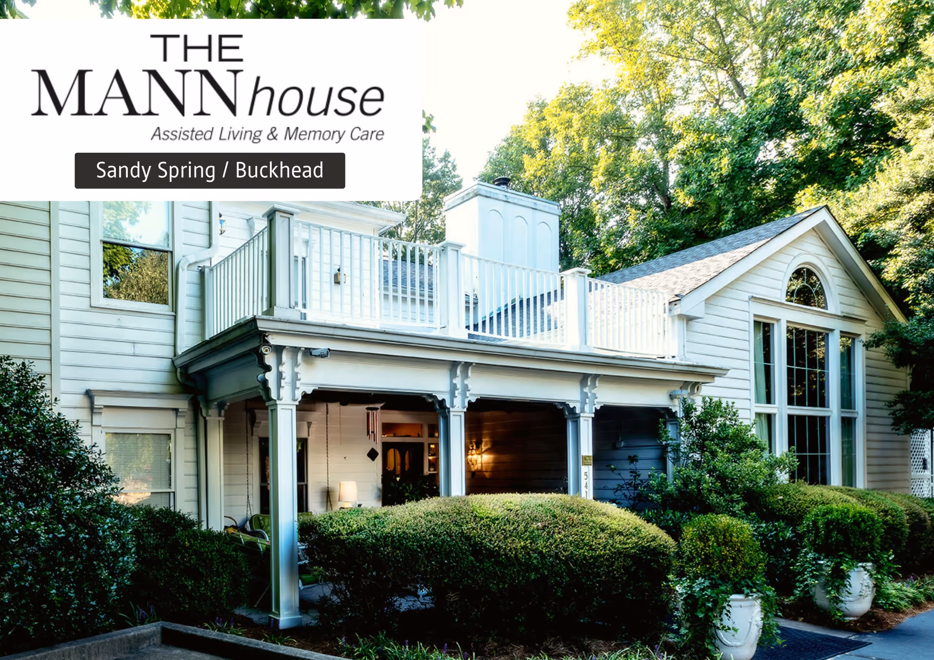 Exterior view of The Mann House Sandy Springs assisted living and memory care facility showing a white building with large windows, a covered porch, and surrounded by green bushes and trees.