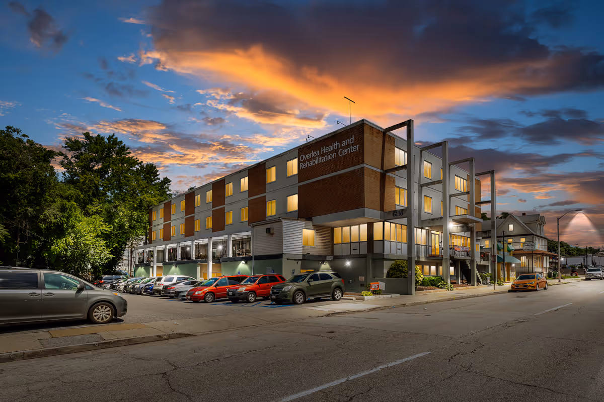 Front exterior of a three-story health and rehabilitation center lit at dusk with a parking lot of cars.